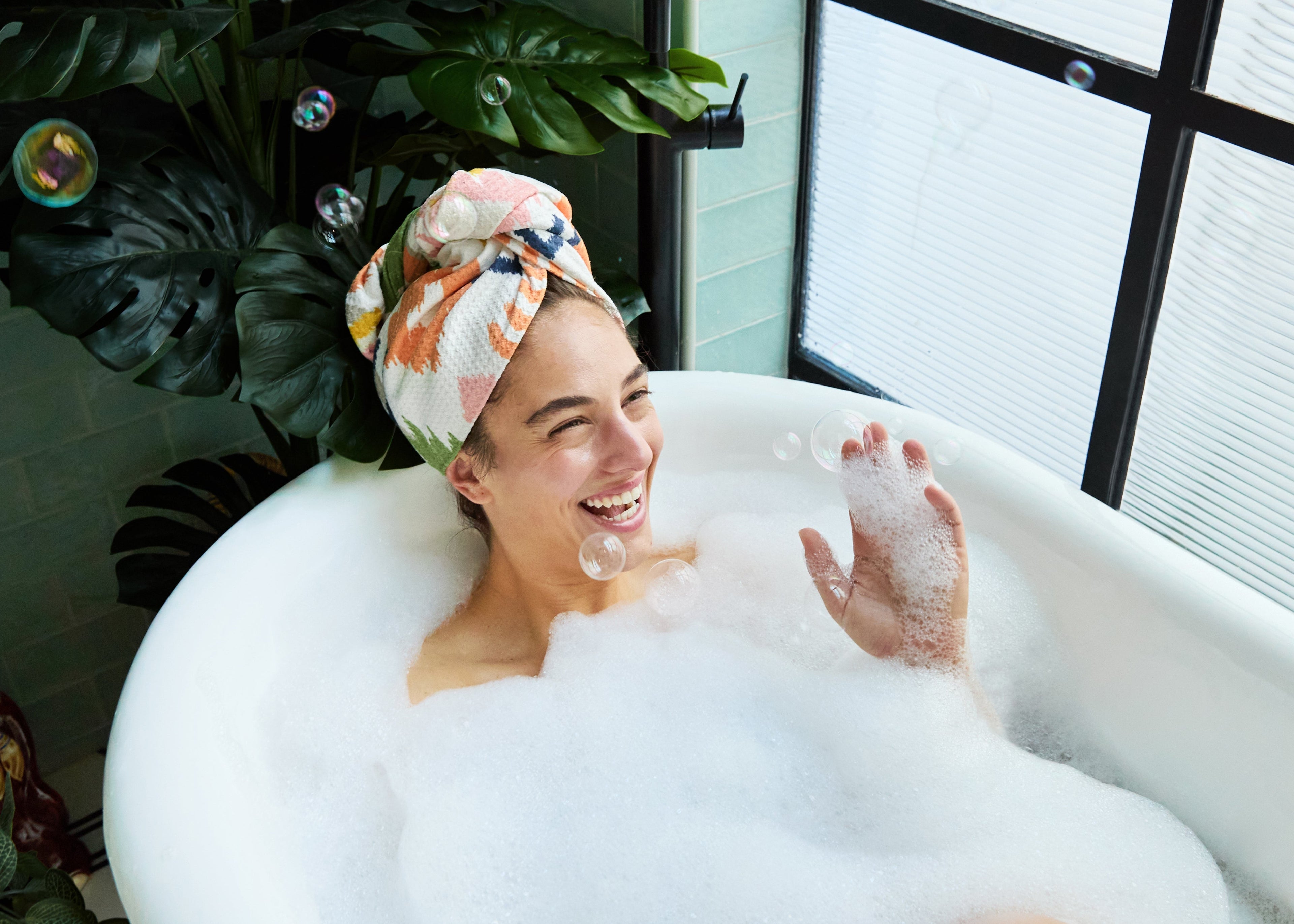 Woman in a bubble bath with a pattern towel on her head, surrounded by plants.