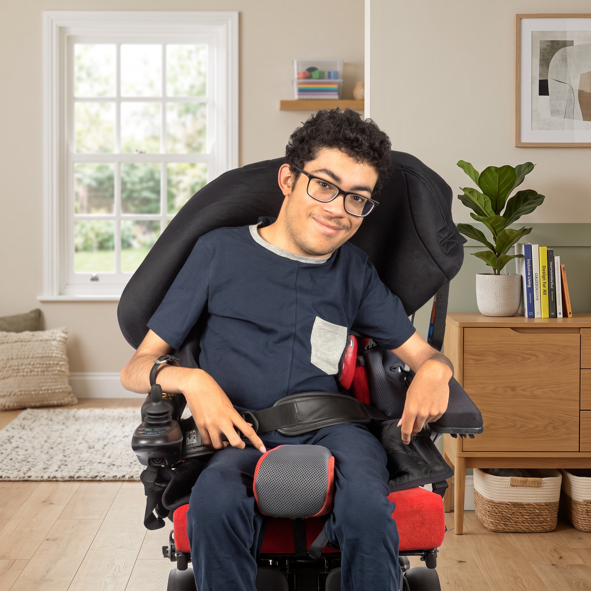 A man wearing glasses sits in a black and red motorized wheelchair in a bright, modern room with a window, wooden cabinet, and potted plant.